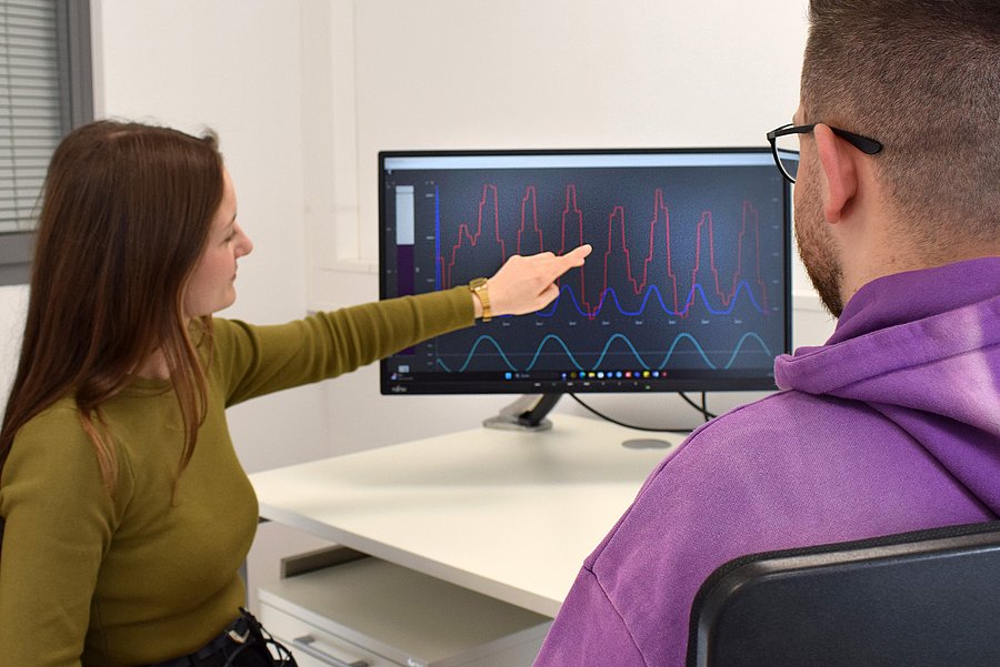 Eine Frau und ein Mann sitzen an einem Schreibtisch in einem modernen Büro. Die Frau trägt ein olivgrünes Oberteil und zeigt mit der Hand auf ein Diagramm auf einem Computerbildschirm. Der Mann, der eine lila Kapuzenjacke trägt, schaut aufmerksam auf den Bildschirm. Das Diagramm zeigt verschiedene Wellenformen in mehreren Farben – eine Analyse von Biofeedback-Daten.