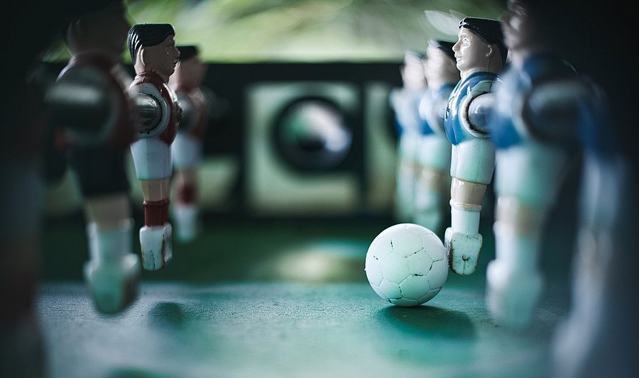 Close-up of a table football game. In the foreground, a white football lies on the green playing field. On the left and right are foosball players in red and blue jerseys, who remain rigidly in their positions. The focus is on the ball, while the figures remain slightly blurred in the background. The picture symbolises structures and fixed roles in sport.