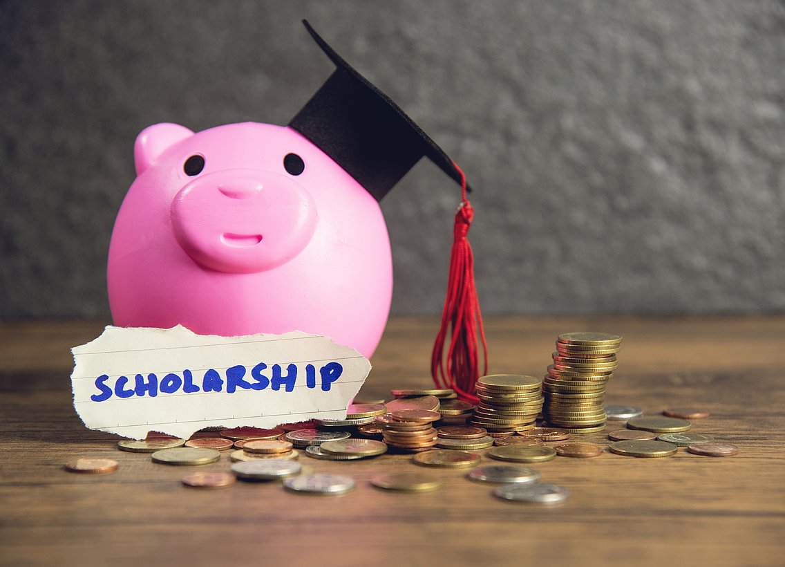 The picture shows a wooden tabletop with a pink-coloured piggy bank on it. This piggy bank is wearing a black university hat and has a note on the front of its belly that says 'Scholarship'. To the right of the piggy bank and in front of the savings slip are euro coins.