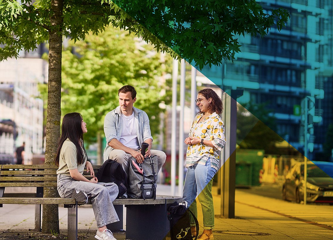 Studierende im Kreis stehend, unterhalten sich vor Unigebäude, auf einer Bank