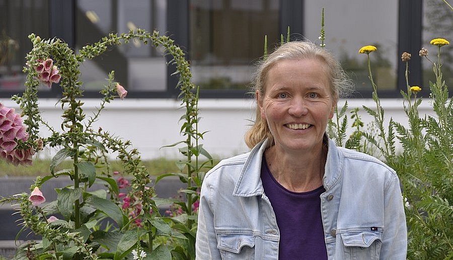 Prof. Gertrud Lohaus steht lächelnd in einer Jeansjacke vor hohen blühenden Pflanzen mit einem Gebäude im Hintergrund.