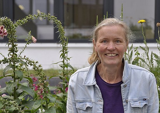 Prof. Gertrud Lohaus steht lächelnd in einer Jeansjacke vor hohen blühenden Pflanzen mit einem Gebäude im Hintergrund.