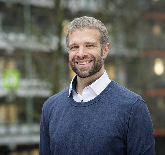 Ein Portrait von Prof. Dr. Philipp Trotter vor der verschwommenen grauen Fassade und den Balkonen der Bergischen Universität Wuppertal 